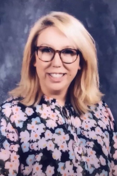 A professional portrait of a smiling woman with blonde hair wearing tortoiseshell glasses and a dark floral blouse with pink and white flowers.