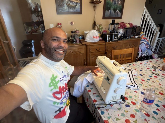 A smiling bald man in a colorful graphic t-shirt sitting at a table, operating a white sewing machine with fabric underneath.