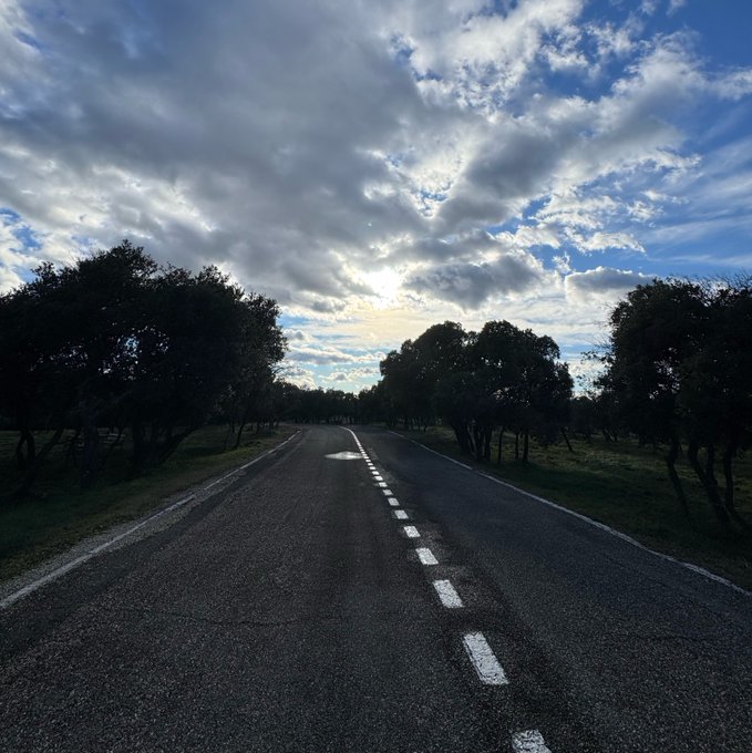 An asphalt road with white dashed lane markings stretches into the distance, flanked by dark green trees under a dramatic sky filled with scattered clouds and sunlight breaking through.