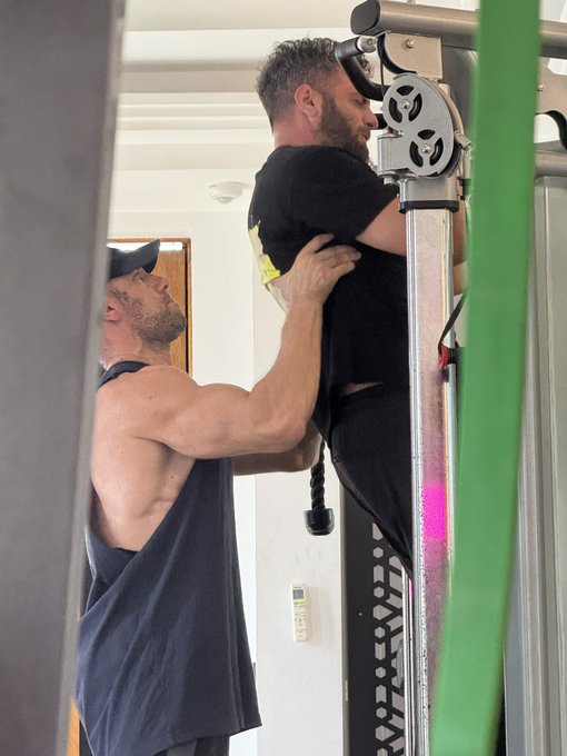 A shirtless man in a black cap assists a bearded man in a black t-shirt at a cable weight machine, guiding him through a workout. The image visually reinforces the post's metaphor of IslandDAO's community strength, mirroring the supportive partnership between Solana figures @solquicks and @thetitanmaker, with the gym setting symbolizing the "strong" foundation of the DAO's ecosystem.