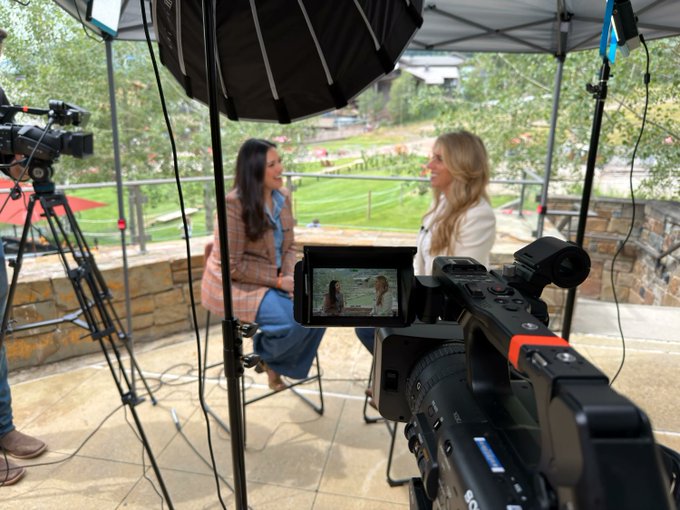 Two women being interviewed outdoors under a tent with professional camera equipment, including a camcorder in the foreground displaying them on its LCD screen.
