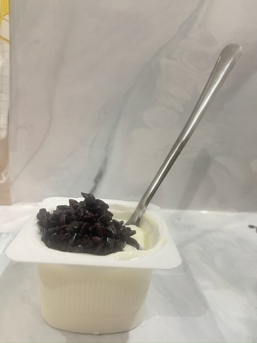 A small white plastic container of yogurt or pudding topped with dark purple/black grains, with a metal spoon sticking out, sitting on a white marble surface.