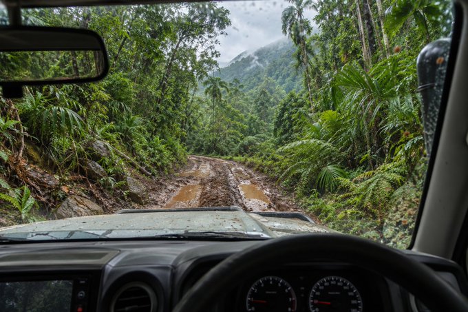 A view from inside a vehicle driving on a muddy jungle road, with lush greenery and mountains visible ahead through the windshield.