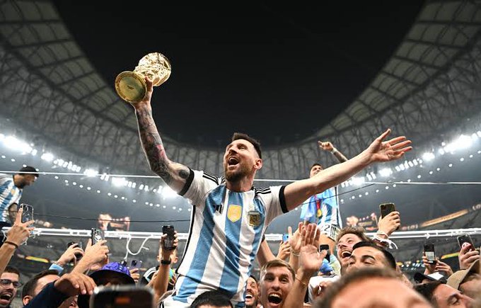 Lionel Messi wearing the Argentina national team jersey, holding up the FIFA World Cup trophy in celebration amidst a crowd of fans in a stadium.