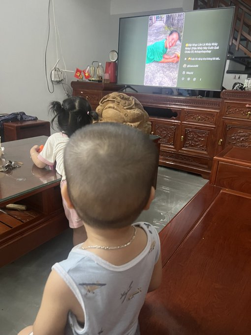 Two young children watching a TikTok video on a large TV in a living room with wooden furniture; the video shows a person lying on the ground.