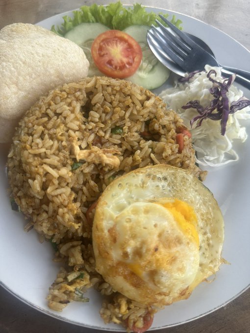 The image shows a plate of food consisting of rice, fried egg, salad, and vegetables such as tomatoes and lettuce. The meal appears to be a healthy option, including a variety of nutrients from the different ingredients. The presence of a fork on the side suggests that this could be a lunch or dinner setting where people are enjoying their meal together.