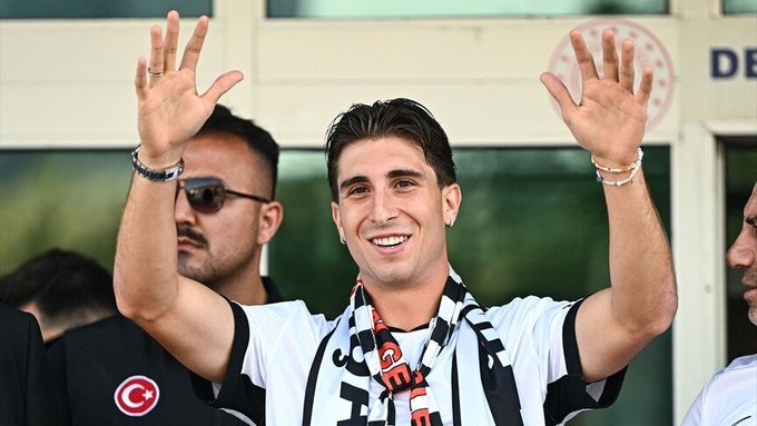 A smiling young man with dark hair waves both hands in the air while wearing a white and black striped Beşiktaş JK jersey and matching scarf, appearing joyful at what looks like a celebratory event, with a man in sunglasses standing behind him near a building marked "DE" and a Turkish flag emblem visible in the background, perfectly matching the caption's sentiment of finding happiness in owning the merchandise despite a loss.