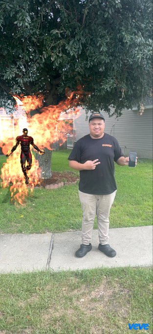 A man wearing an 'xStocks' t-shirt stands on a sidewalk holding a phone, with an augmented reality fiery figure and explosion visible in the grass behind him, branded with a 'veve' watermark.