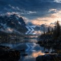 A scenic landscape photograph showing a mountain range and pine trees reflected in a calm lake at sunset, with a '>' shaped logo overlay in the center.