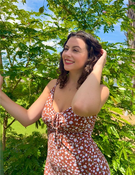 A woman in a floral dress smiles brightly while standing under lush green foliage, embodying the post's message about allowing yourself to smile and embrace joy. Her genuine, radiant expression captures the essence of the reminder to find happiness in simple moments.