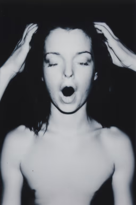 A black-and-white close-up photograph captures a woman with her eyes closed and mouth open, her hands raised near her head.
