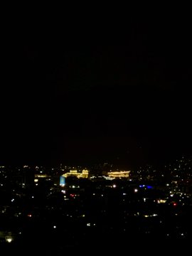A nighttime cityscape view showing scattered illuminated buildings and streetlights against a dark sky.