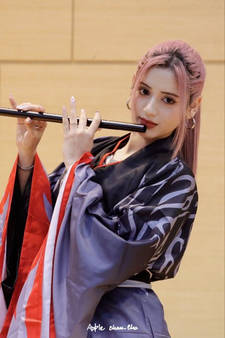 A woman with pink hair styled up poses in a red, black, and purple traditional-style robe, holding a black flute-like object; text reads 'Apple_chan_elle'.