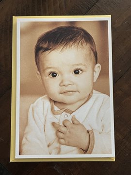 A sepia-toned greeting card featuring a vintage portrait photograph of a baby wearing a light-colored shirt, placed on a dark wooden surface.