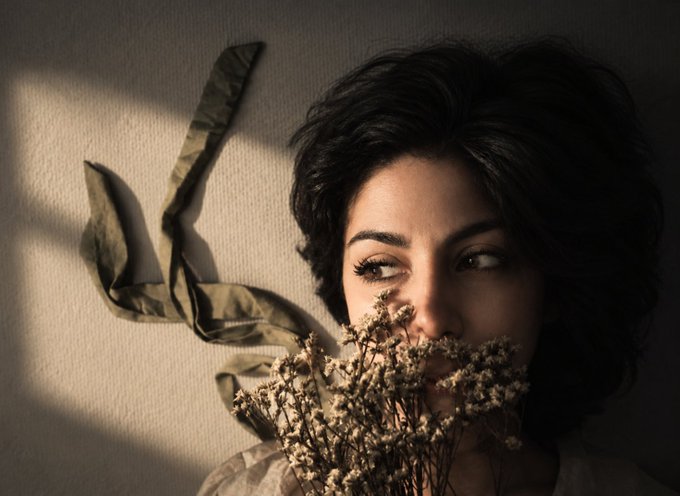 A woman with short dark hair is partially obscured by a bouquet of dried flowers, with sunlight casting window pane shadows and illuminating a draped green ribbon on the wall.