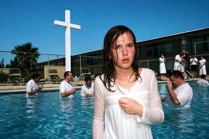 A film still of a young woman in a wet white dress standing in a swimming pool, with a large white cross and people in white robes visible in the background.
