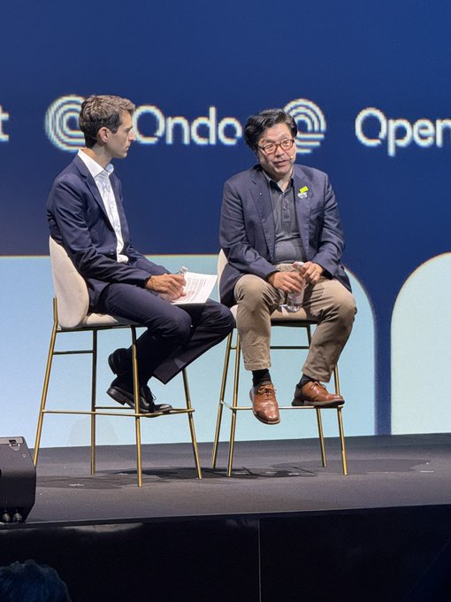 Two men in business attire sit on a stage at what appears to be an 'Ondo' conference, one holding papers while the other gestures as he speaks.