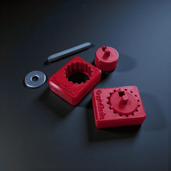 Red 3D-printed objects including square and circular mechanical parts with gear-like teeth, along with a metal washer and a gray rod, are arranged on a dark surface.
