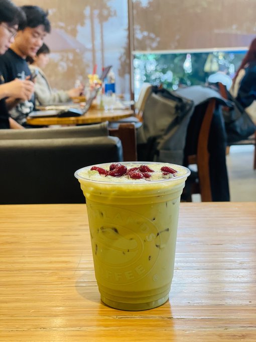 A green matcha drink topped with red goji berries sits in a clear plastic cup branded 'Caffè Bene' on a wooden table, with people visible working on laptops in the background of a café.