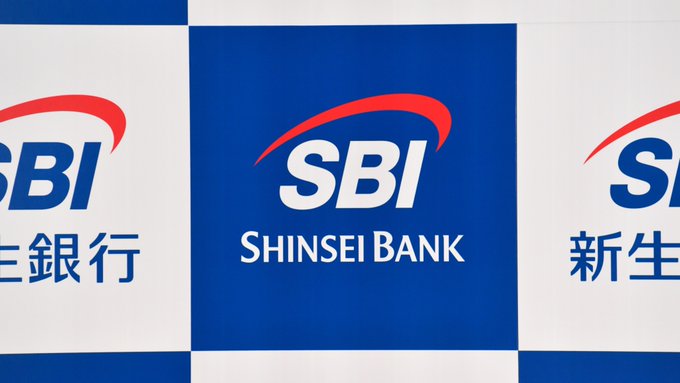 The image features a blue and white sign for Shinsei Bank that says "SBI" at the top of the sign, followed by "Bank". Below this, there are three lines of text written in Japanese, which appear to be related to cryptocurrency or digital currency. The text reads: "Japan's SBI Shinsei Bank will let customers convert part of their deposit interest into Bitcoin, Ethereum, or XRP. The pilot launches on June 10." This suggests that the bank is exploring ways to incorporate and support digital currencies in its services for its customers.