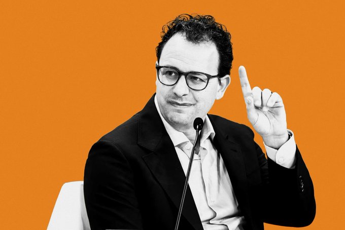 A man in a black suit and white shirt, wearing black-rimmed glasses, sits at a microphone against an orange background, raising his index finger as if making an important announcement—visually echoing the breaking news about Anthropic's rumored new Claude AI model and its potentially record-breaking $2 trillion IPO in November.