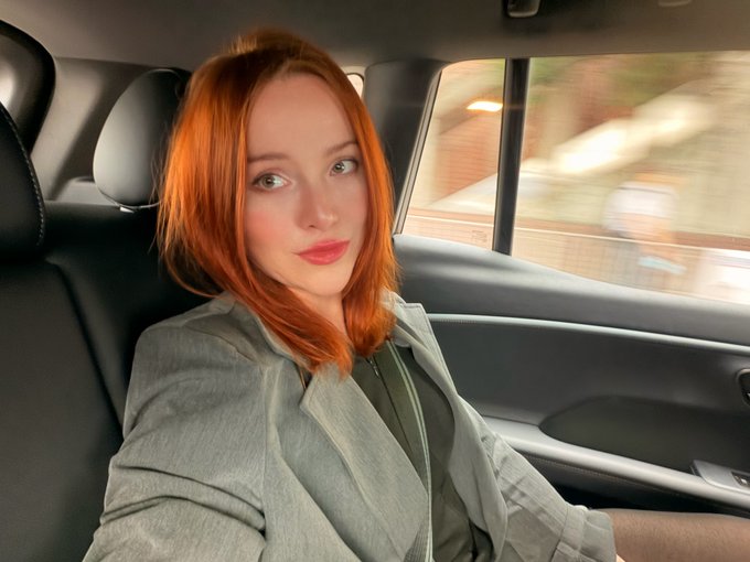 A selfie of a woman with bright orange/red hair wearing a gray blazer, sitting in the back seat of a moving car.