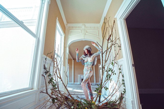 A body-painted figure wearing a twig hat poses in a sunlit hallway framed by branches, visually representing the "Washington State Photographic portrait" described in the text as available on Objkt.