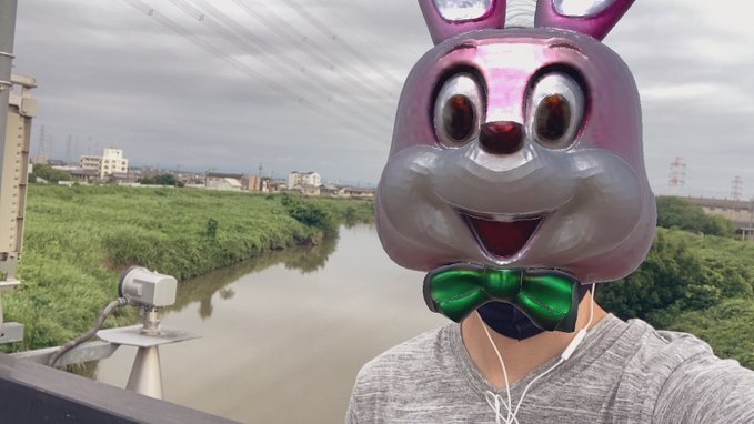 A person wearing a purple bunny mask and a green bow tie stands on a bridge over a river, wearing white earphones and a gray shirt, with a small camera mounted on the railing. The image relates to the post about a 42km running challenge on #STEPN, as the person appears to be preparing to start or document their run after the typhoon has passed.