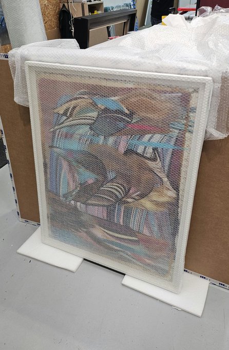 An abstract painting wrapped in bubble wrap is propped against cardboard boxes, resting on white foam packaging blocks in what appears to be a storage or retail area.