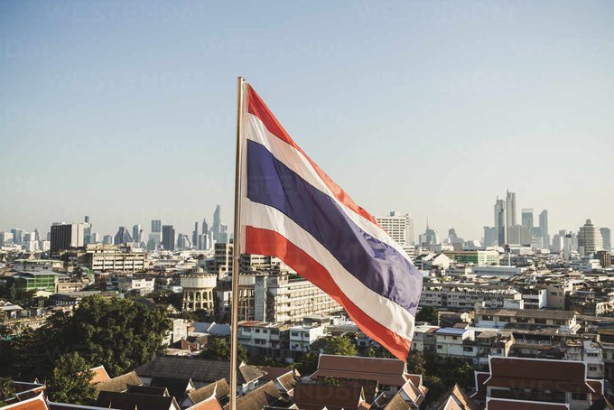 The Thai flag (red, white, and blue stripes) flies prominently in the foreground with the modern skyline of Bangkok, Thailand visible in the background under a hazy sky.