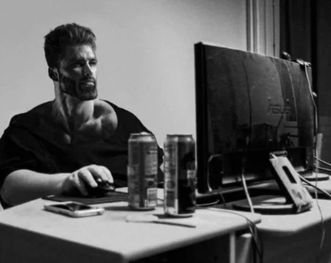 A black and white photo of a man at a desk using a computer with ASUS monitor, with energy drinks and a phone on the desk.