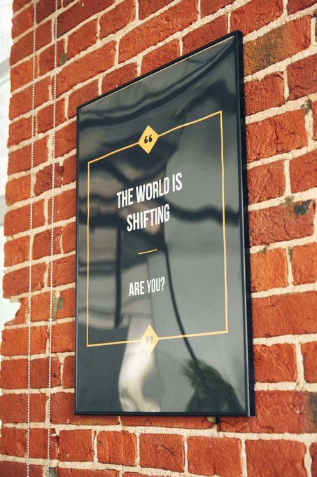A framed black and yellow motivational poster mounted on a red brick wall that reads "THE WORLD IS SHIFTING — ARE YOU?".