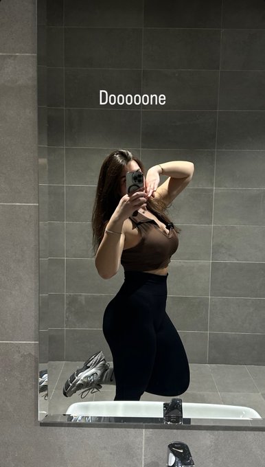 A mirror selfie of a woman with long brown hair wearing a brown crop top and black leggings, posing in a grey-tiled room, with the text 'Dooooone' overlaid at the top.