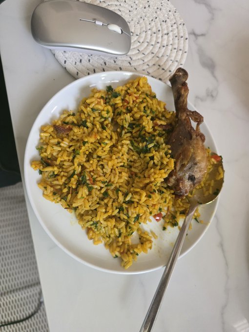 A white plate containing yellow seasoned rice mixed with herbs, served alongside a piece of cooked chicken on the bone, with a spoon and a silver computer mouse resting on a woven coaster nearby.