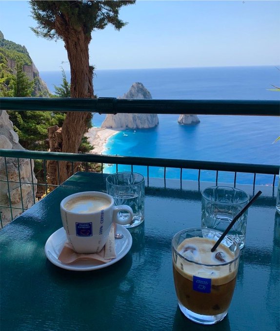 A coffee cup and iced coffee sit on a green table overlooking a stunning turquoise cove with rocky outcrops at the Mediterranean coastline. The "100% Capri" branded cups serve as a picturesque backdrop for promoting BeldexCoin's privacy-focused crypto ecosystem, juxtaposing the tranquil seaside view with the post's tech-forward message about $BDX.