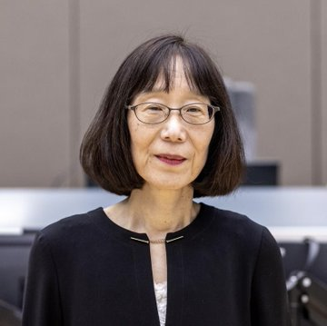 A woman with short dark hair and glasses, wearing a black cardigan, stands in what appears to be an office or institutional setting. The image accompanies a post discussing global political persecution, though no specific text, names, or identifying details are visible in the photo itself.