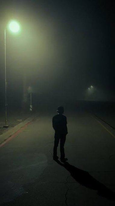 A moody, atmospheric photograph of a lone silhouetted figure standing on a foggy street under a glowing streetlamp at night.