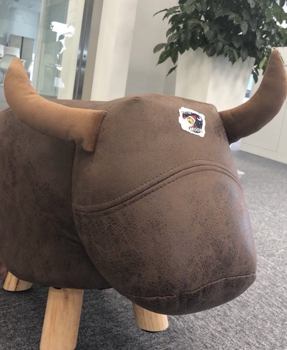 A photo of a brown bull-shaped stool with large horns and a 'MOON' label on its side, displayed indoors near a potted plant on a carpeted floor.
