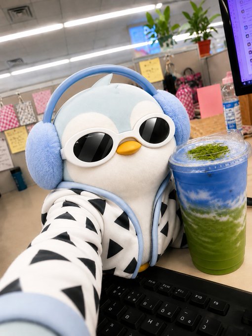 A cute plush penguin toy wearing blue headphones and sunglasses sits at a desk next to a keyboard and a green-blue layered matcha drink in a plastic cup.