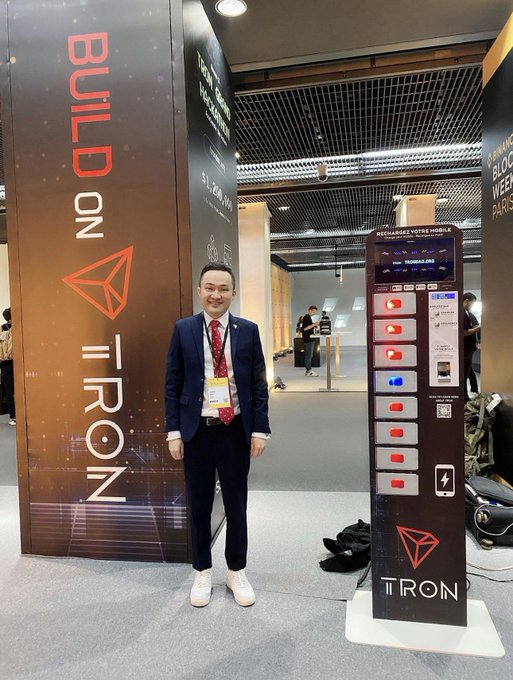 A man in a blue suit and red tie stands at a TRON booth at what appears to be a tech conference, with 'BUILD ON TRON' signage and a recharging station visible beside him.