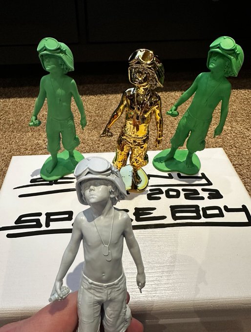 A collection of small 3D-printed figurines resembling Space Boy, with versions in green, gold, and gray plastic, displayed on a base labeled '2023 Space Boy'.