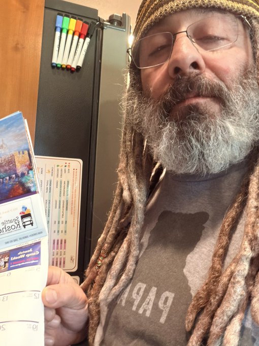 A selfie of a bearded man with dreadlocks, glasses, and a knit cap, standing in front of a black refrigerator with colorful markers on top, holding a grocery store receipt and coupons for Casey's and Eureka points.