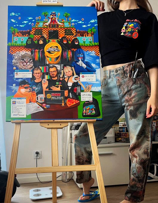A person wearing a black graphic t-shirt and tie-dye jeans poses next to a colorful Nintendo-themed canvas artwork on an easel, featuring characters and a 'Freshly Picked' tomato soup reference.