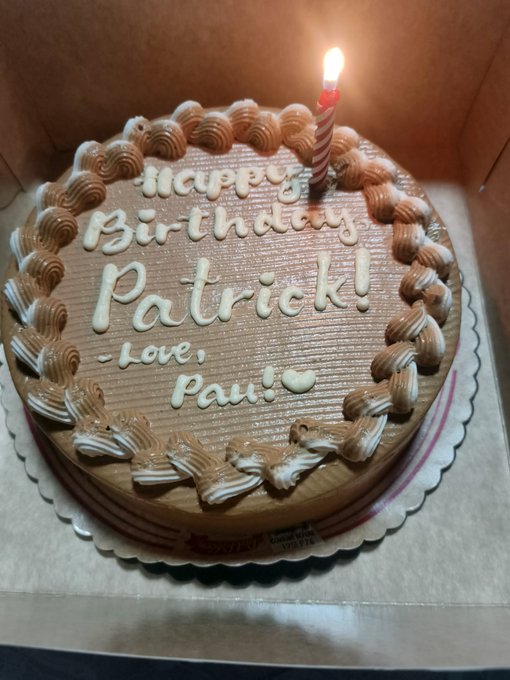 A chocolate cake in a box has piped "Happy Birthday Patrick! Love, Paul!" with a lit candle on top.