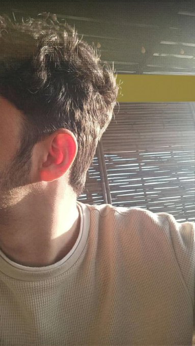 A side-profile selfie of a man with dark hair and stubble, his ear illuminated in red sunlight, wearing a cream-colored waffle-knit shirt with horizontal window blinds in the background.