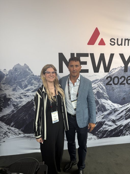 A man and woman in business attire posing together in front of a summit backdrop reading 'NEW Y... 2026' with a mountain scene.