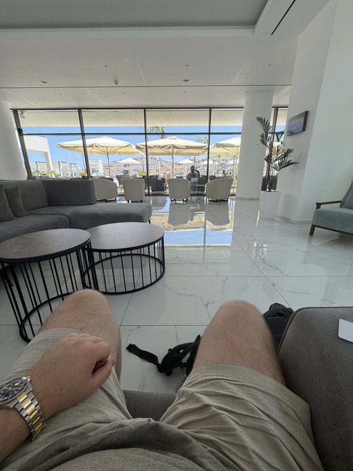 The image shows a first-person perspective of someone relaxing in a modern hotel lounge while waiting for their car to the airport, with their legs visible in the foreground and a rooftop terrace with umbrellas, seating, and a cityscape view in the background.