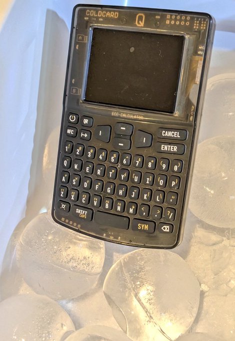 A transparent Coldcard Q cryptocurrency hardware wallet with a full QWERTY keyboard and screen, resting on a surface with cylindrical containers.
