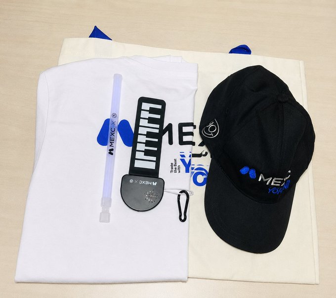 A flat-lay of promotional merchandise including a white MEXC t-shirt, a black MEXC cap, a tote bag, a pen, and a carabiner.