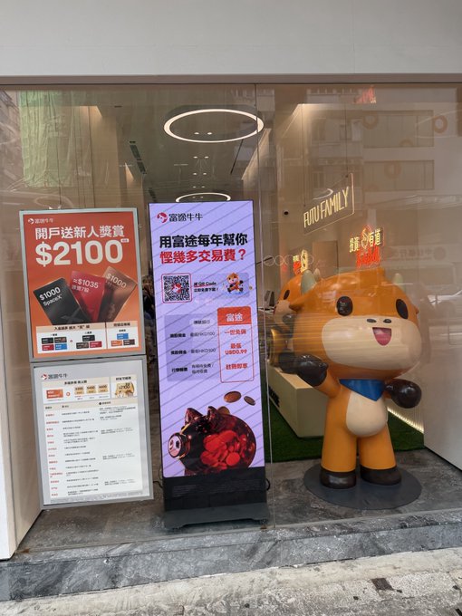 The entrance of a Richufamily (富途牛牛) store featuring a large orange cartoon bull mascot, alongside promotional banners in Chinese advertising new user bonuses of $2100 and crypto trading fees.